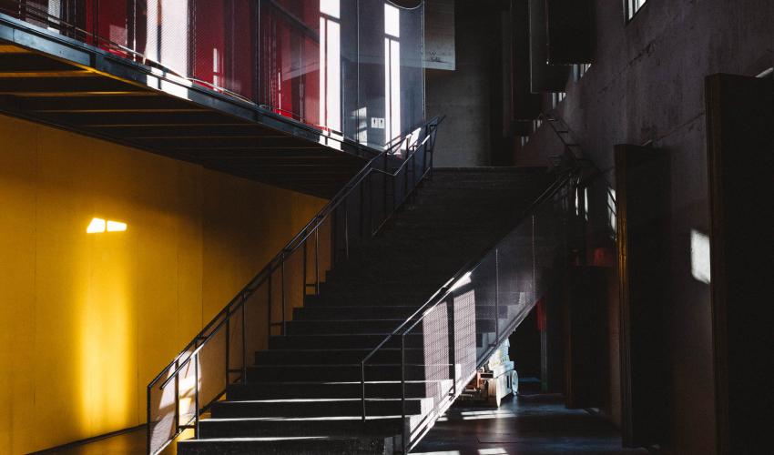 Imagen vertical de un foyer o hall principal de color negro. Hay una escalera negra, un panel amarillo e ingreso de luz natural. 